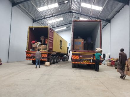 Two large trucks in a warehouse with people standing around.